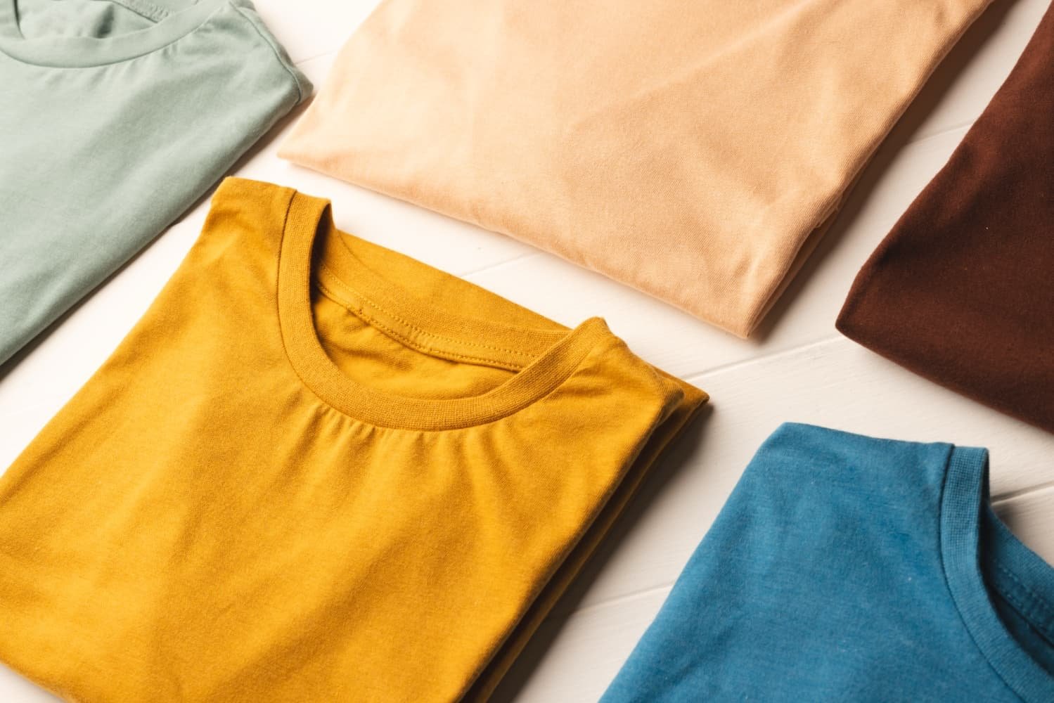 Close-up top view of stacked folded sweatshirts in mustard yellow, navy blue, cream, and beige with ribbed collars.