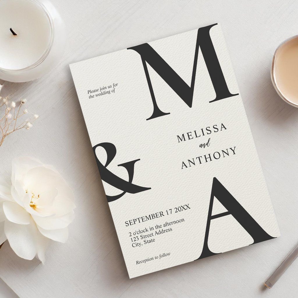 Elegant black and white wedding invitation featuring large serif initials "M & A" on textured paper with a white flower and candle nearby.