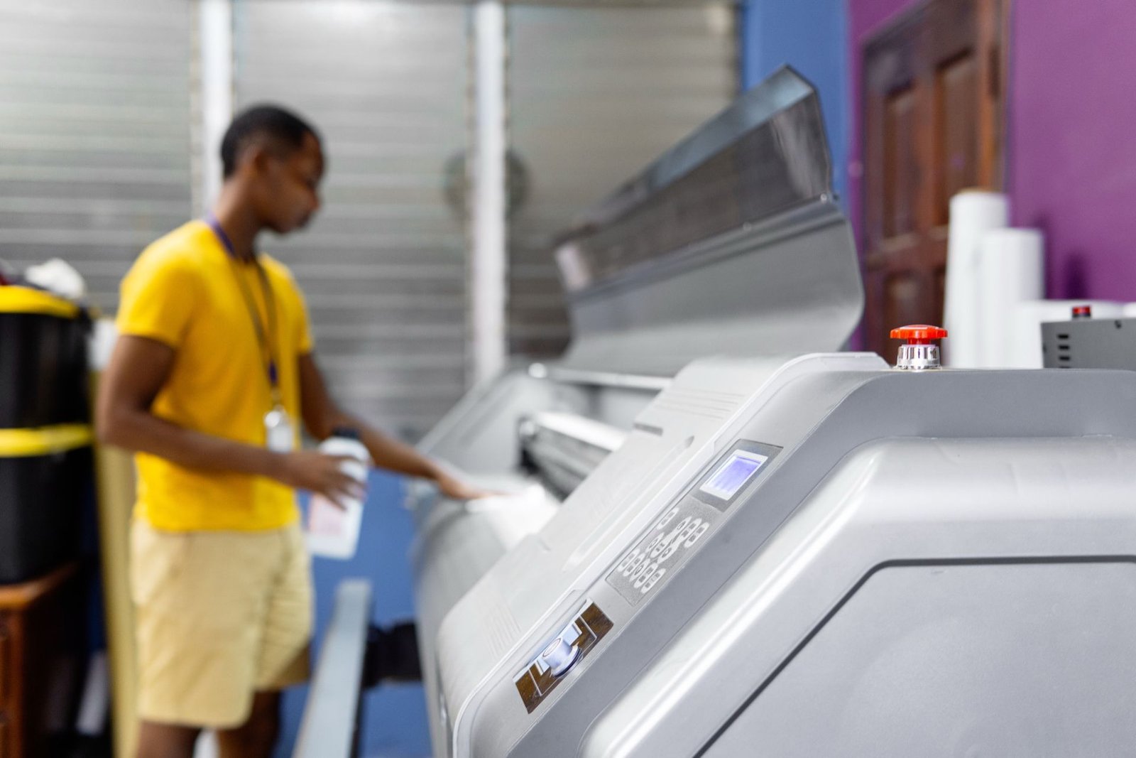 Blurred worker in a yellow shirt operating a large-format grey commercial printer in a print shop.